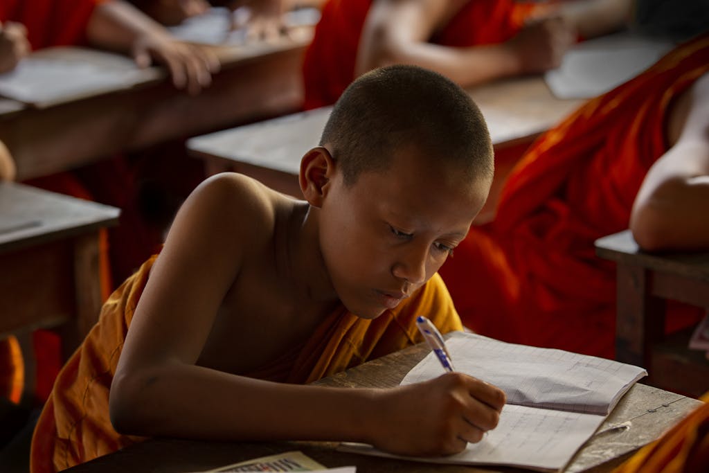 A young monk wearing orange robes attentively writing in a classroom setting, highlighting education.